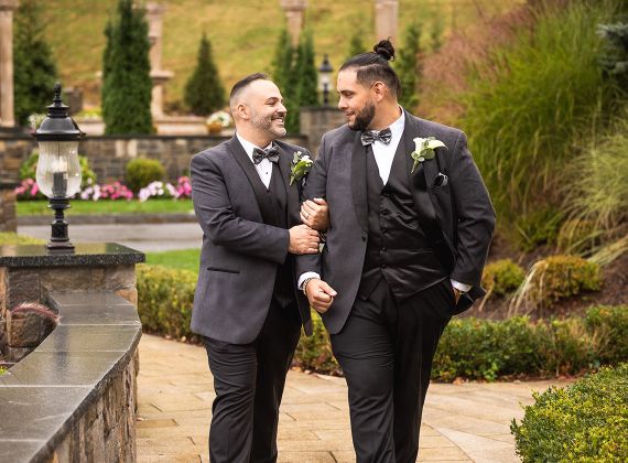 Two grooms in elegant tuxedos smiling at each other in a garden with greenery and flowers.